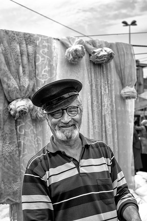 The Curtain Seller's Smile - A monochromatic study captures a fleeting moment of genuine warmth and character, set against a backdrop of layered textures